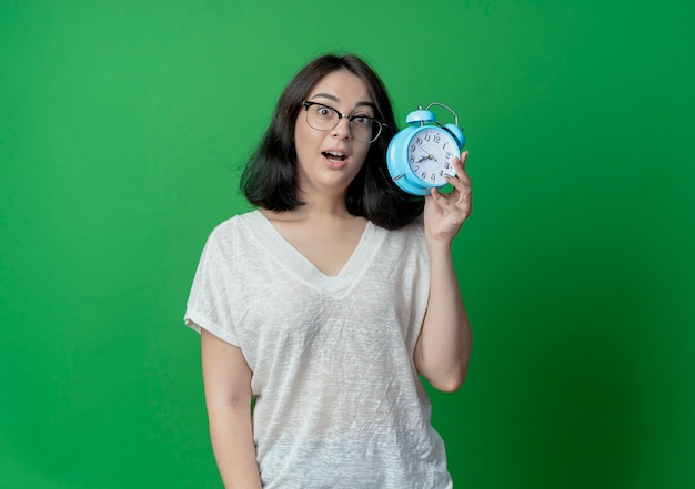 Impressed young pretty woman wearing glasses holding alarm clock