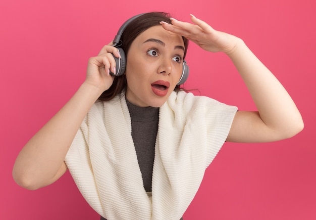 Impressed young pretty woman wearing and grabbing headphones keeping hand near forehead looking at side into distance