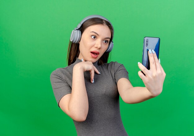Впечатленная молодая красивая женщина в наушниках, держащая и смотрящая на мобильный телефон и держащая руку на груди, изолированную на зеленом фоне с копией пространства