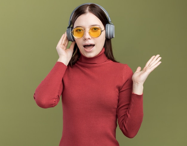 Impressed young pretty woman wearing sunglasses and headphones touching headphones showing empty hand