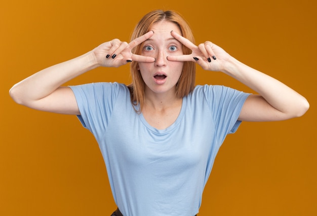 Foto gratuita giovane ragazza rossa impressionata dello zenzero con le lentiggini che gesturing il segno di vittoria attraverso le dita isolate sulla parete arancione con lo spazio della copia