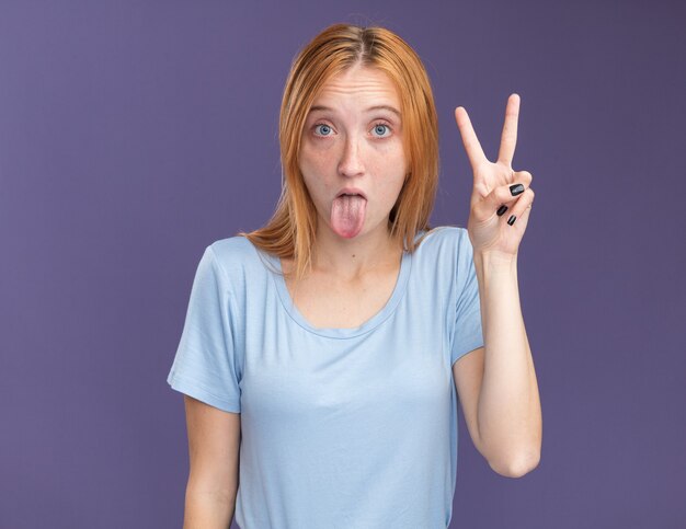 Impressed young redhead ginger girl with freckles stucks out tongue and gestures victory sign isolated on purple wall with copy space