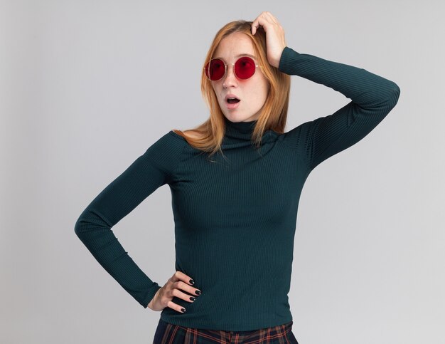 Impressed young redhead ginger girl with freckles in sun glasses puts hand on head and looks at side isolated on white wall with copy space