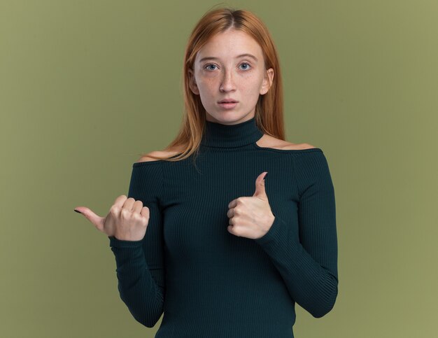 Impressed young redhead ginger girl with freckles thumbs up and points at side isolated on olive green wall with copy space