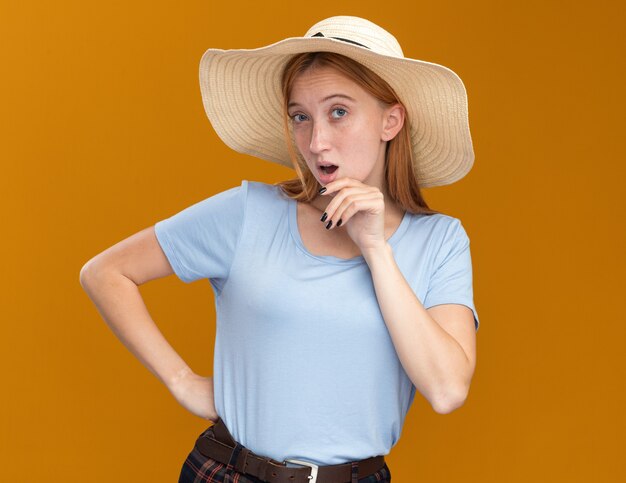 Impressed young redhead ginger girl with freckles wearing beach hat holding chin and looking at camera on orange