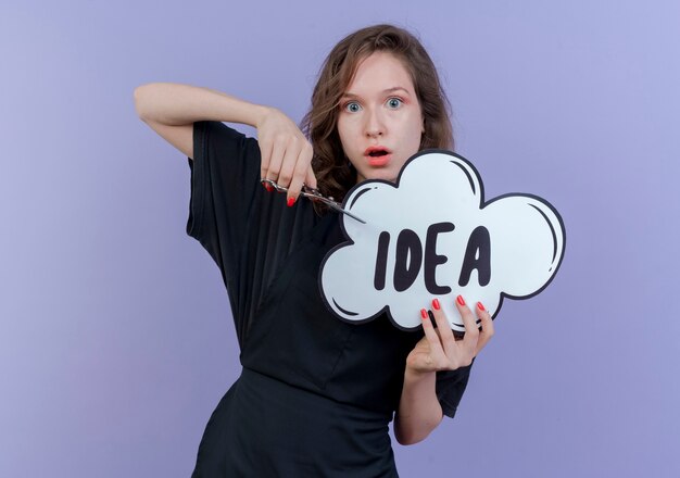 Impressed young slavic female barber wearing uniform holding idea bubble and scissors isolated on purple background