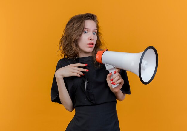 Impressed young slavic female barber wearing uniform holding speaker keeping hand in air isolated on orange background with copy space