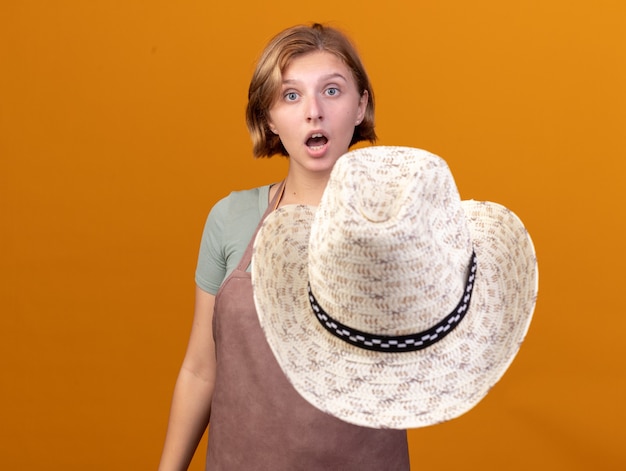 Impressed young slavic female gardener holding gardening hat and looking at camera on orange