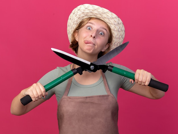 Free photo impressed young slavic female gardener wearing gardening hat stucks out tongue and holds gardening scissors isolated on pink wall with copy space