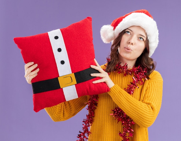 Impressed young slavic girl with santa hat and with garland around neck holding decorated pillow looking up isolated on purple background with copy space