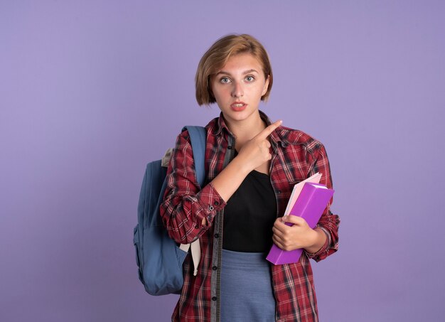 Impressed young slavic student girl wearing backpack holds book and notebook points at side 