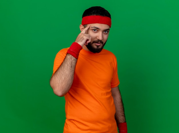 Impressed young sporty man wearing headband and wristband putting finger on temple isolated on green background