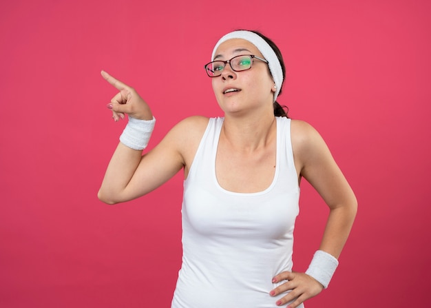 Impressed young sporty woman in optical glasses wearing headband and wristbands puts hand on waist and points at side isolated on pink wall