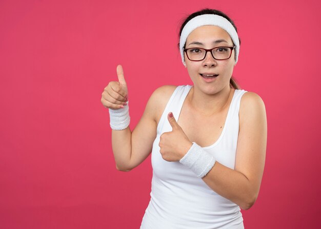 Впечатленная молодая спортивная женщина в оптических очках, носящая повязку на голову и браслеты, большие пальцы руки вверх двух рук, изолированные на розовой стене