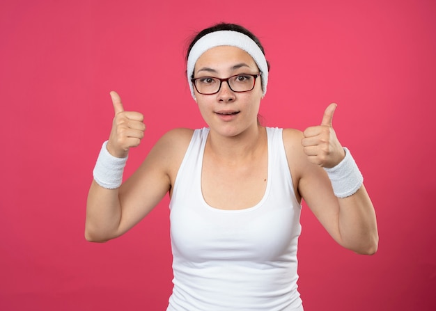 Впечатленная молодая спортивная женщина в оптических очках, носящая повязку на голову и браслеты, показывает палец вверх двумя руками, изолированными на розовой стене