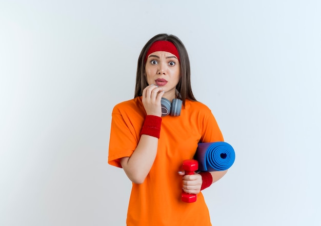Впечатленная молодая спортивная женщина с повязкой на голову, браслетами и наушниками на шее, держащая гантель с ковриком для йоги, касаясь подбородка, выглядящая изолированной