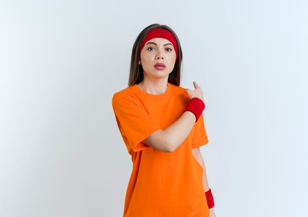 Impressed young sporty woman wearing headband and wristbands looking pointing behind isolated