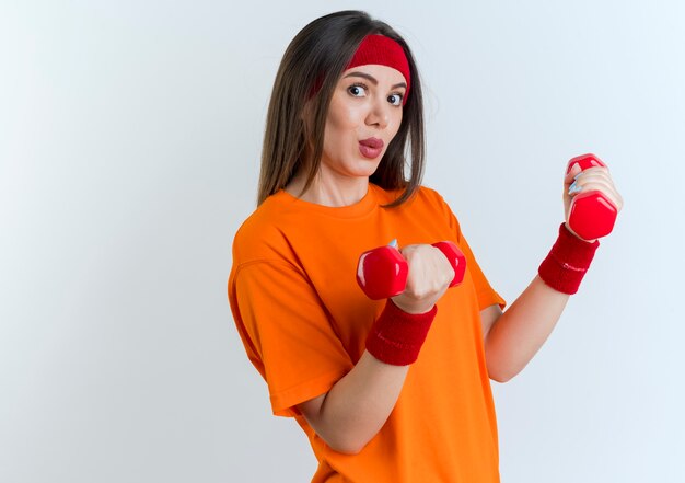 Impressed young sporty woman wearing headband and wristbands standing in profile view  holding dumbbells isolated on white wall with copy space