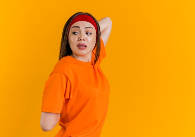 Impressed young sporty woman wearing headband and wristbands standing in profile view looking at side keeping hands behind back exercising isolated on orange wall with copy space