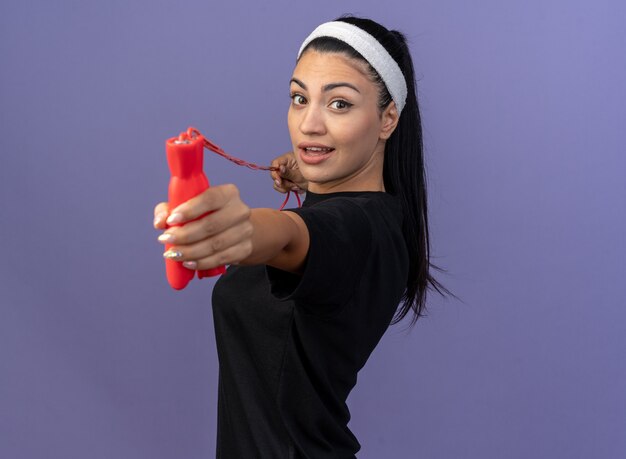 Впечатленная молодая спортивная женщина с повязкой на голову и браслетами, стоящая в профиль, тянет скакалку и вытягивает ее вперед, глядя вперед, изолированную на фиолетовой стене