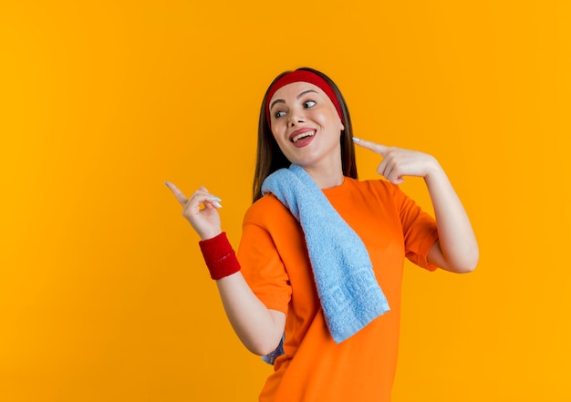 Впечатленная молодая спортивная женщина, носящая повязку на голову и браслеты с полотенцем на плече, смотрит и указывает на сторону, изолированную на оранжевой стене с копией пространства