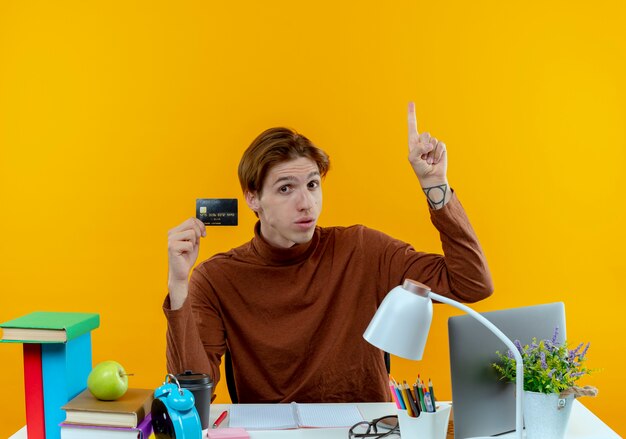 impressed young student boy sitting at desk with school tools holding credit card and points at up