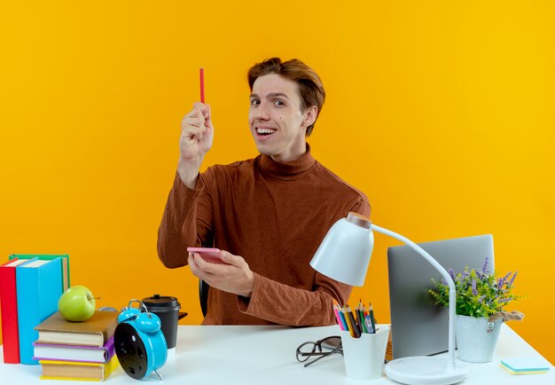 Impressed young student boy sitting at desk with school tools holding notebook and raising pen 