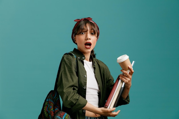 Impressed young student girl wearing bandana and backpack holding note pads and paper coffee cup looking at camera isolated on blue background