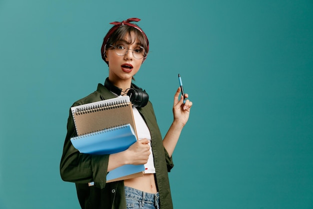 Impressed young student girl wearing bandana glasses and headphones around neck standing in profile view holding large note pads looking at camera pointing up with pen with copy space