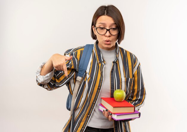 Impressed young student girl wearing glasses and back bag holding books with apple on it pointing and looking down isolated on white