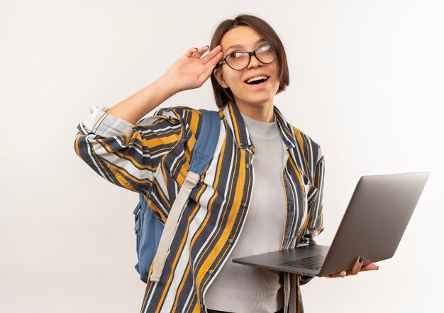 Impressed young student girl wearing glasses and back bag holding laptop looking at side putting hand on temple isolated on white