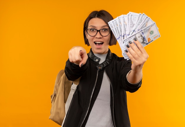 Impressed young student girl wearing glasses and back bag holding money and pointing at front isolated on orange