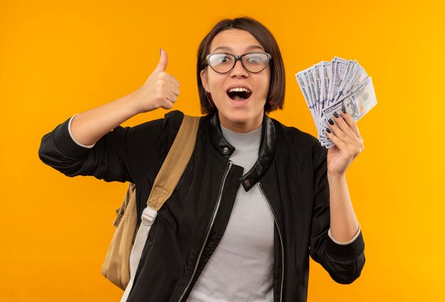 Impressed young student girl wearing glasses and back bag holding money showing thumb up isolated on orange