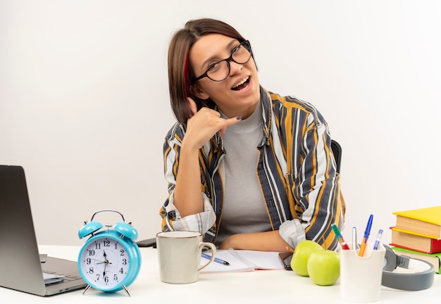 Impressed young student girl wearing glasses sitting at desk doing call sign isolated on white