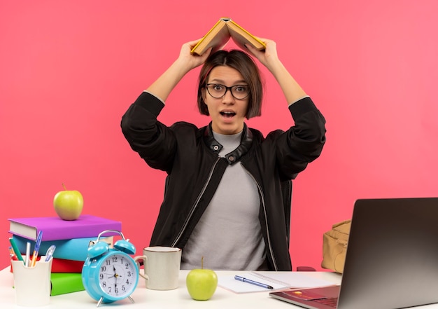 Impressed young student girl wearing glasses sitting at desk doing homework keeping book above head isolated on pink