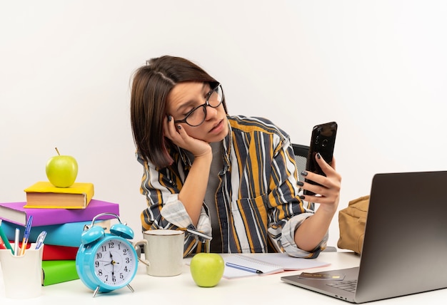 Impressed young student girl wearing glasses sitting at desk holding and looking at mobile phone isolated on white