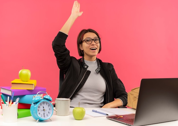 Impressed young student girl wearing glasses sitting at desk raising hand isolated on pink