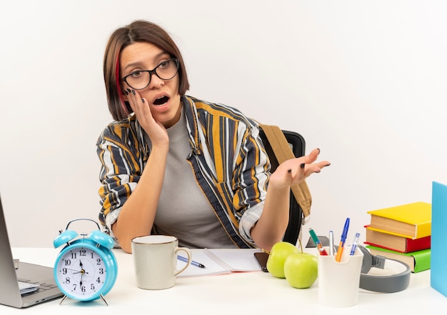 Free photo impressed young student girl wearing glasses sitting at desk showing empty hand with hand on face looking at side isolated on white