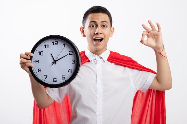 Impressed young superhero boy in red cape holding clock looking at camera doing ok sign isolated on white background