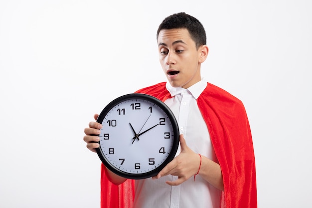 Impressed young superhero boy in red cape holding and looking at clock isolated on white background with copy space