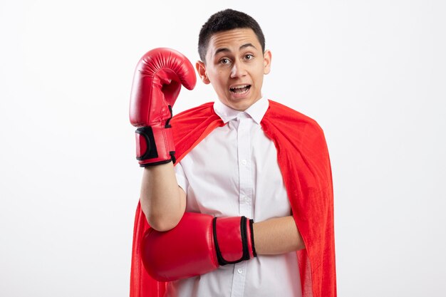 Impressed young superhero boy in red cape wearing box gloves looking at camera keeping hand in air isolated on white background with copy space