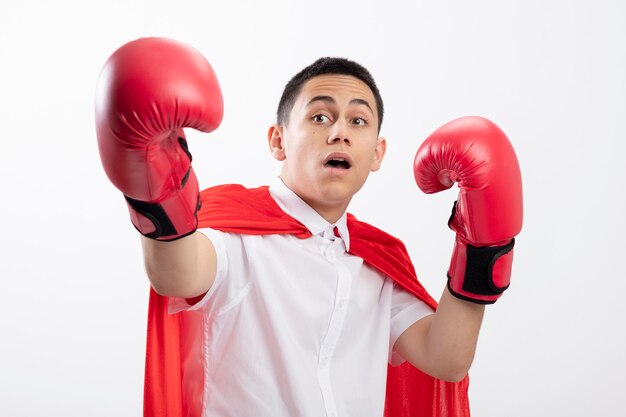 Impressed young superhero boy in red cape wearing box gloves looking at side keeping hand in air stretching another hand out towards camera isolated on white background