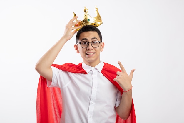 Impressed young superhero boy in red cape wearing glasses looking at camera grabbing crown pointing at it isolated on white background with copy space