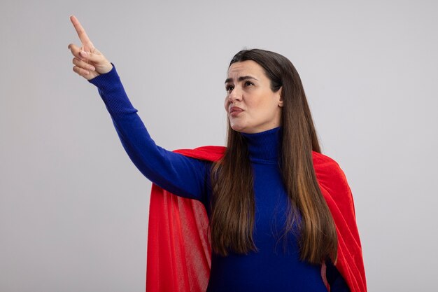 Impressed young superhero girl looking and points at side isolated on white background