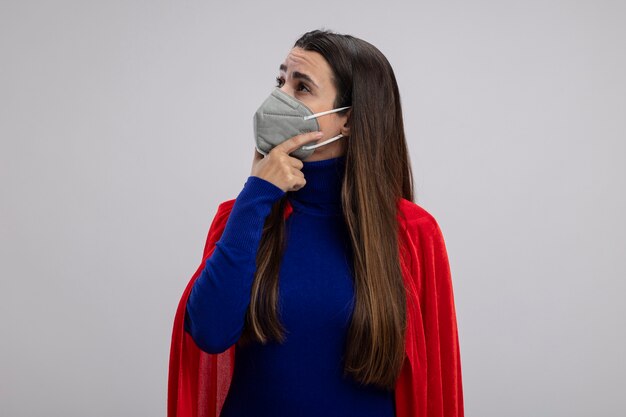 Impressed young superhero girl looking at side wearing medical mask putting hand under chin isolated on white