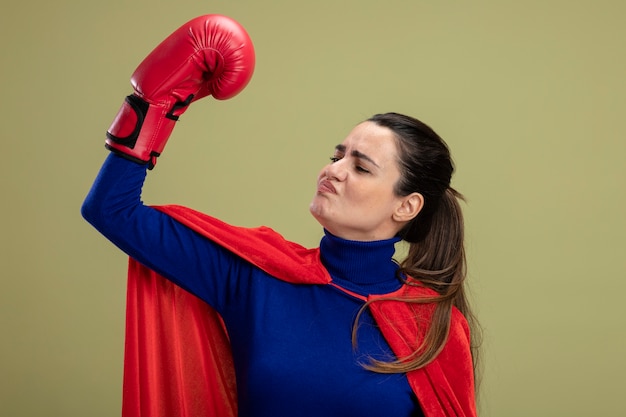 オリーブグリーンで隔離の強いジェスチャーを示すボクシンググローブを身に着けている感動の若いスーパーヒーローの女の子