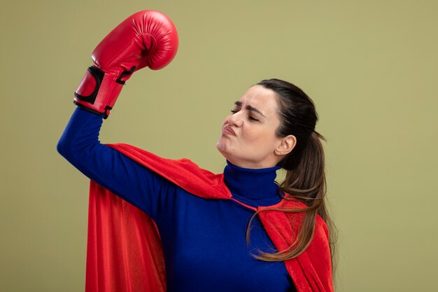 Impressed young superhero girl wearing boxing gloves showing strong gesture isolated on olive green