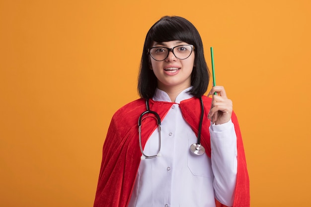 Впечатленная молодая девушка-супергерой в стетоскопе с медицинским халатом и плащом с очками, поднимающими карандаш