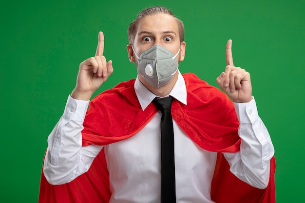 Free photo impressed young superhero guy wearing medical mask and tie points at up isolated on green background