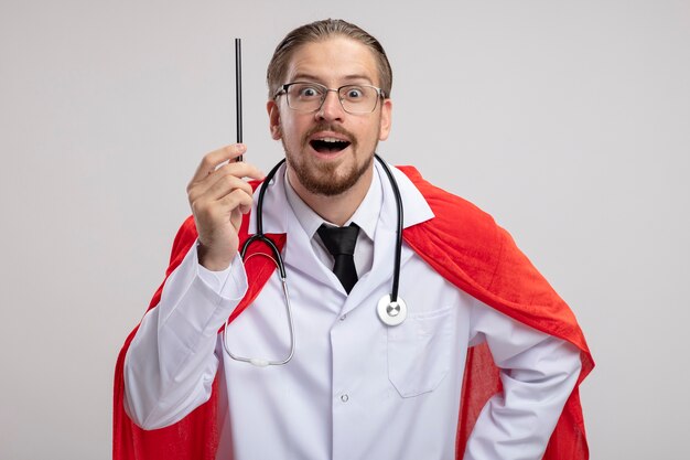 Impressed young superhero guy wearing medical robe with stethoscope and glasses holding pencil